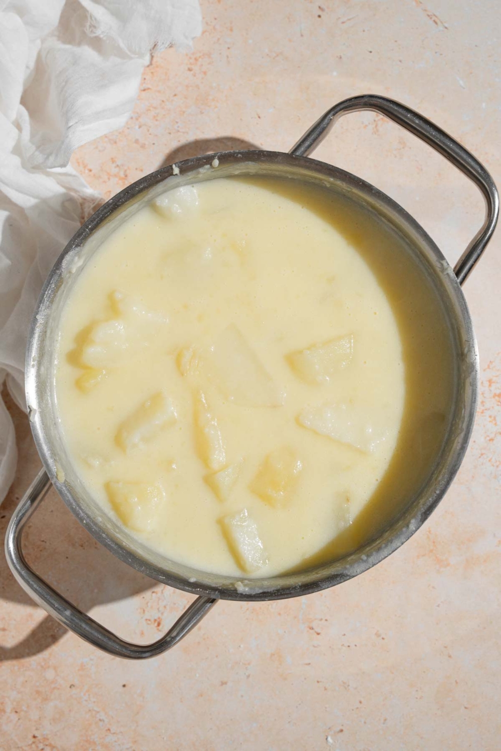 Potatoes cooking in a milk and flour slurry in a pot. The pot is on a tan counter with a white cloth napkin.