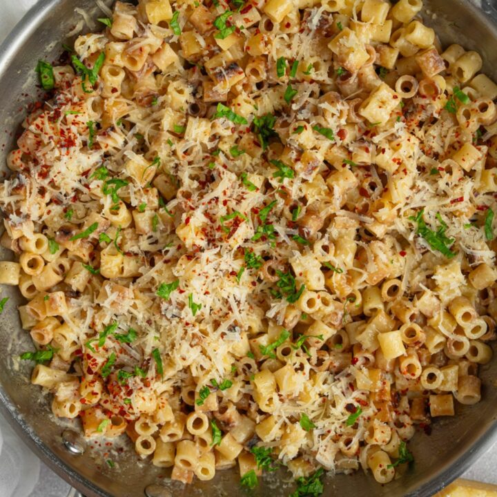 A pan with ditalini pasta garnished with fresh parsley and grated cheese. The pan is on a white counter with a ramekin of fresh parsley, a wedge of parmesan cheese, and ramekin of red pepper flakes.