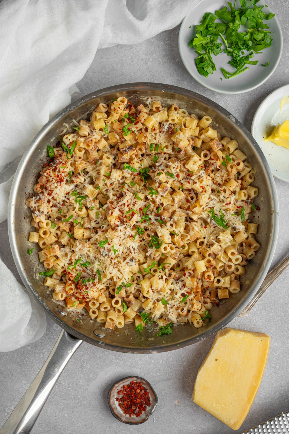 A pan with ditalini pasta garnished with fresh parsley and grated cheese. The pan is on a white counter with a ramekin of fresh parsley, a wedge of parmesan cheese, and ramekin of red pepper flakes.