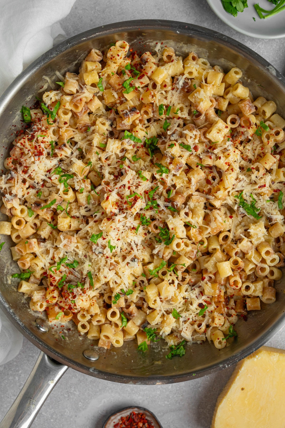 A pan with ditalini pasta garnished with fresh parsley and grated cheese. The pan is on a white counter with a ramekin of fresh parsley, a wedge of parmesan cheese, and ramekin of red pepper flakes.