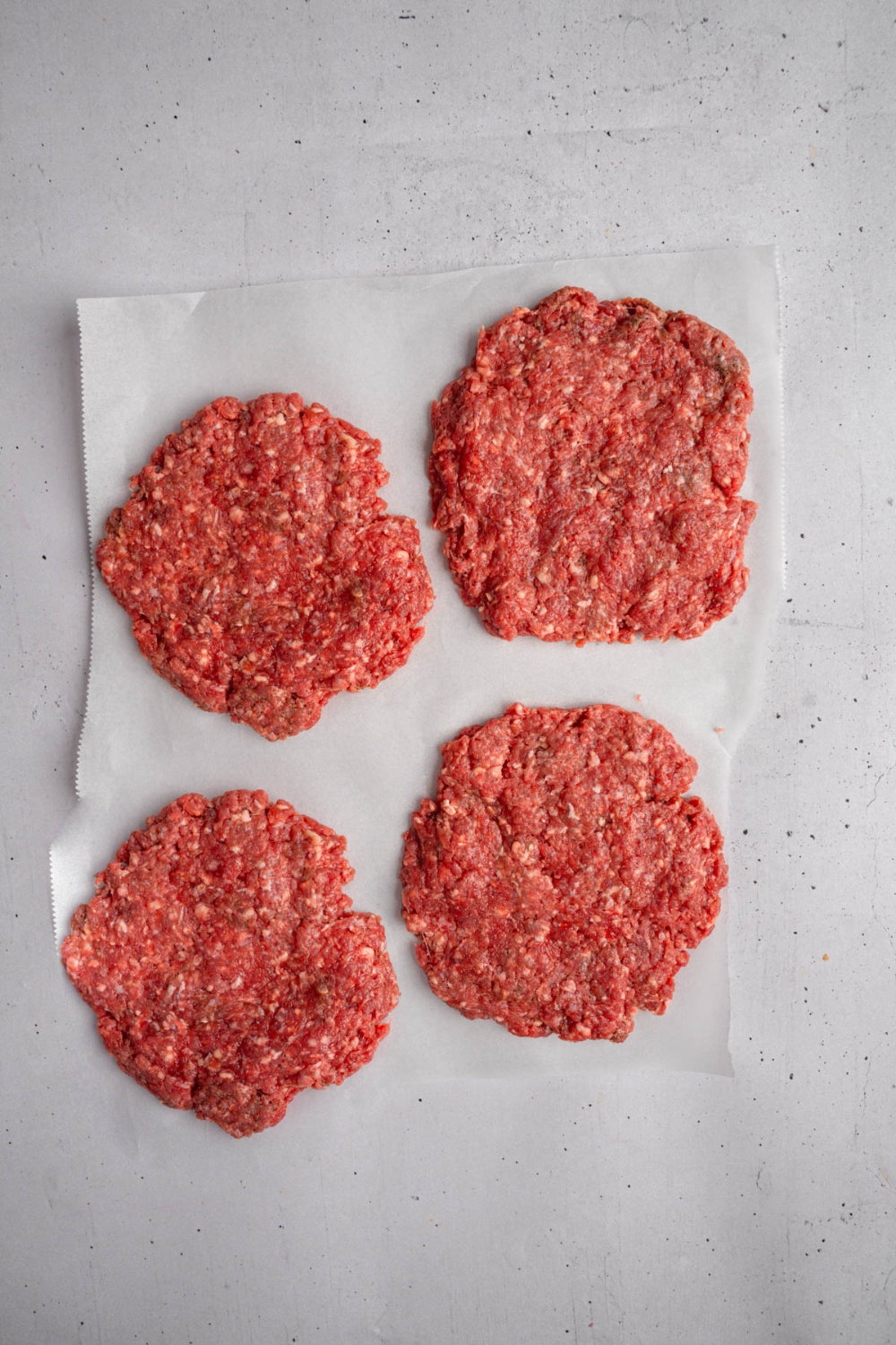 Four uncooked burger patties on a piece of parchment paper. The paper is on a white counter.