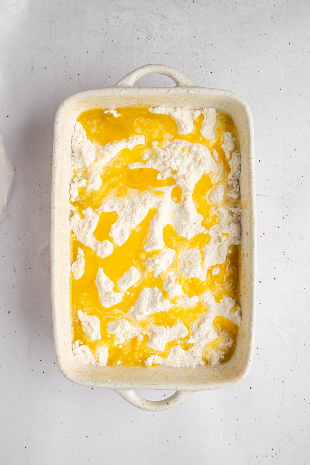 A baking dish with pecan pie mixture topped with cake mix and melted butter. The dish is on a white counter.