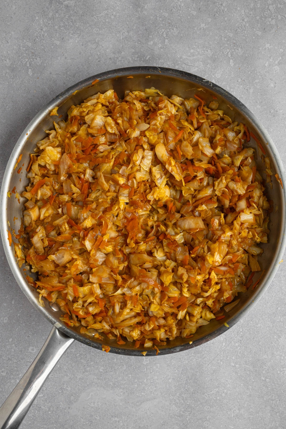 A stainless steel frying pan with chopped carrots, cabbage, onion, and garlic cooking in oil and sauces. The pan is on a white counter.