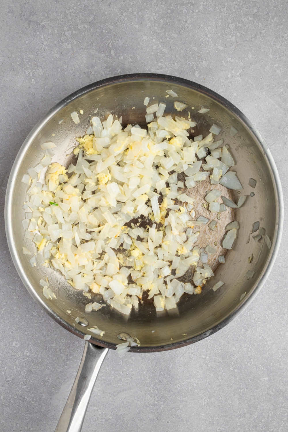 A stainless steel frying pan with chopped white onion and garlic frying in oil.