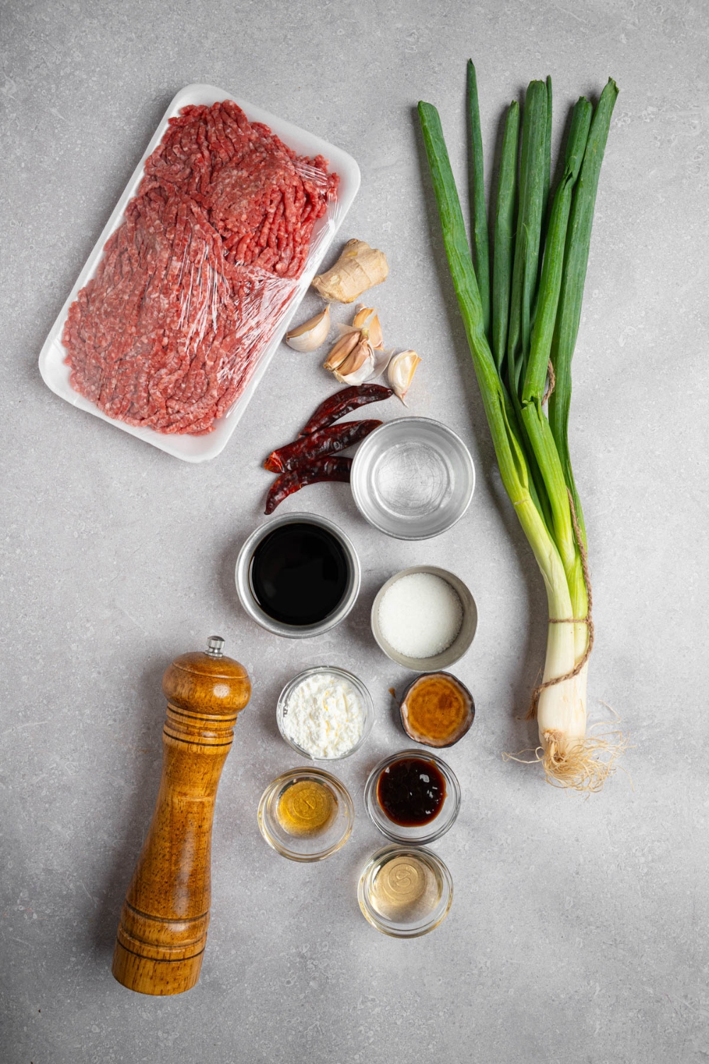 An overhead shot of several bowls in various sizes containing ingredients to make Mongolian ground beef including ground beef, green onions, dried chilis, garlic, ginger, soy sauce, oil, cornstarch, and more.