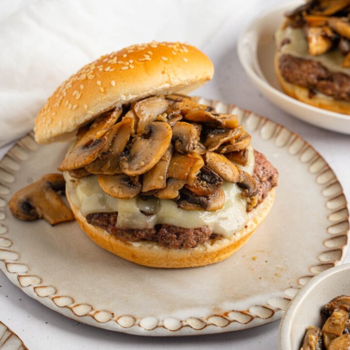 An open mushroom swiss burger on a ceramic plate. The plate is on a white counter with an additional burger and a white cloth napkin.