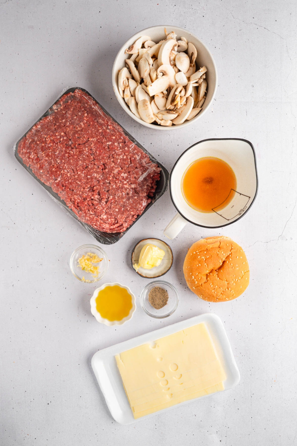 An overhead shot of several bowls in various sizes containing ingredients to make mushroom swiss burgers including ground beef, sliced mushrooms, worcestershire sauce, garlic, swiss cheese, butter, beef broth, and seasonings.