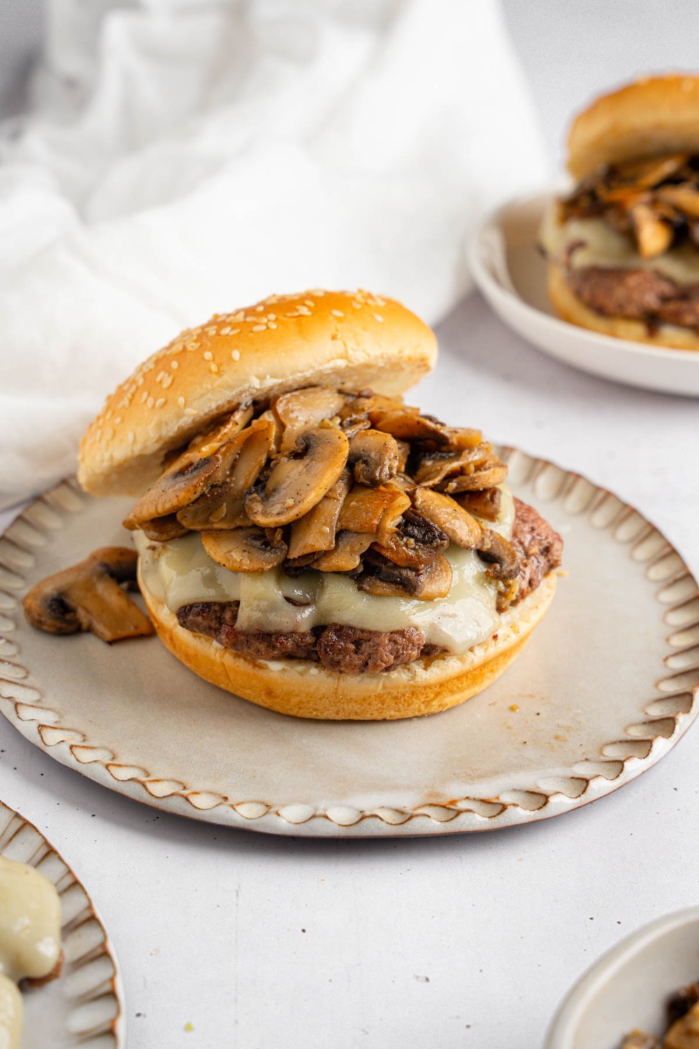 An open mushroom swiss burger on a ceramic plate. The plate is on a white counter with an additional burger and a white cloth napkin.