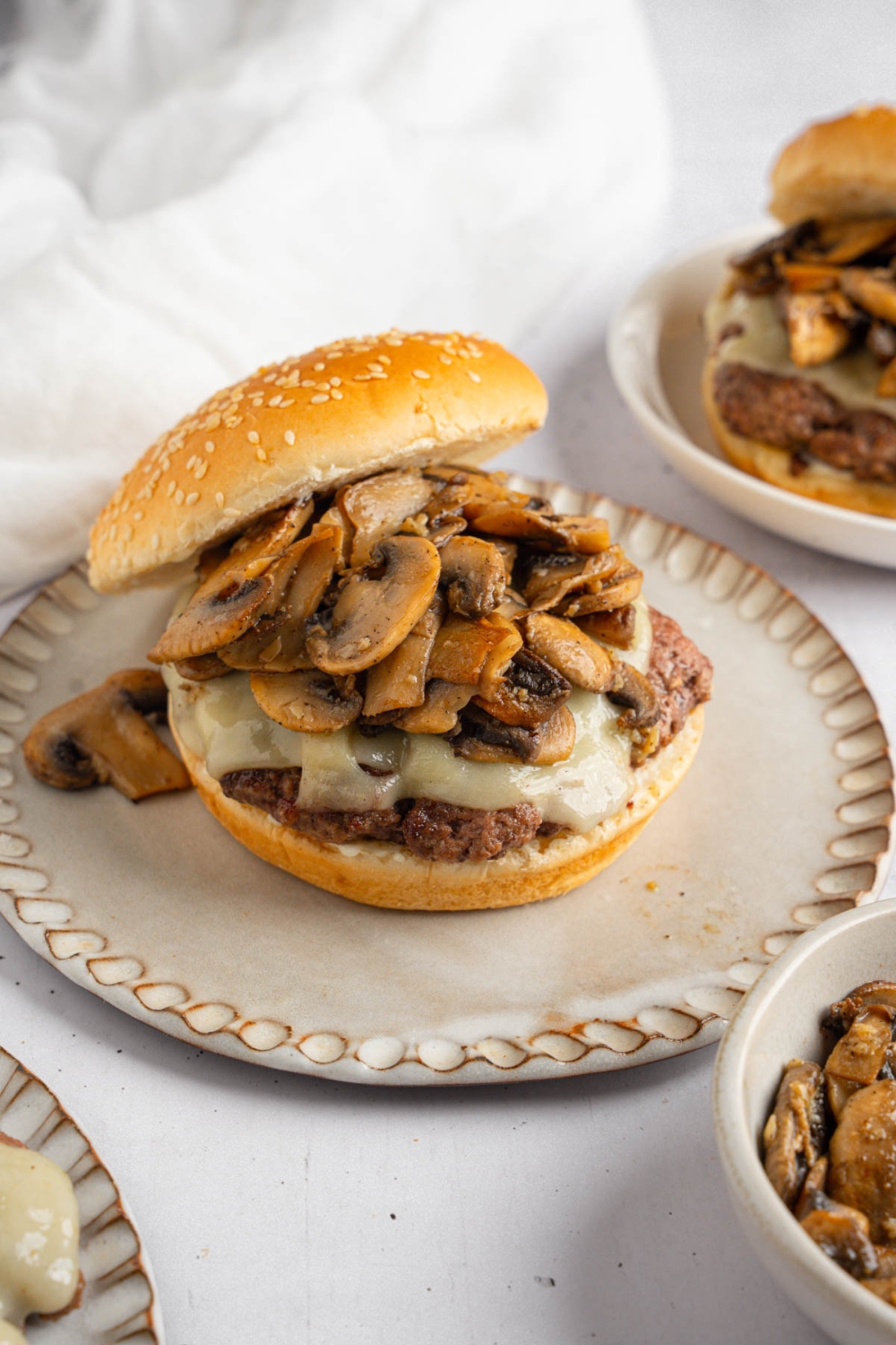 An open mushroom swiss burger on a ceramic plate. The plate is on a white counter with an additional burger and a white cloth napkin.