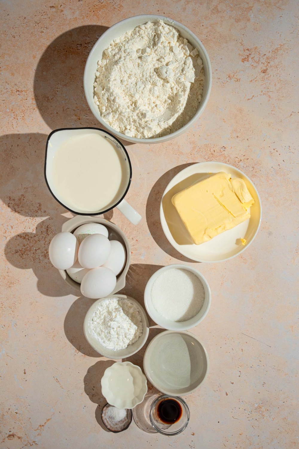 An overhead shot of several bowls in various sizes with ingredients to make Napoleon cake including flour, eggs, sugar, salt, butter, vanilla, vinegar, water, milk, and more.