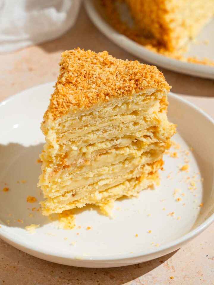 A slice of Napoleon cake on a white plate. The plate is on a counter with a white cloth napkin and Napoleon cake on a plate.