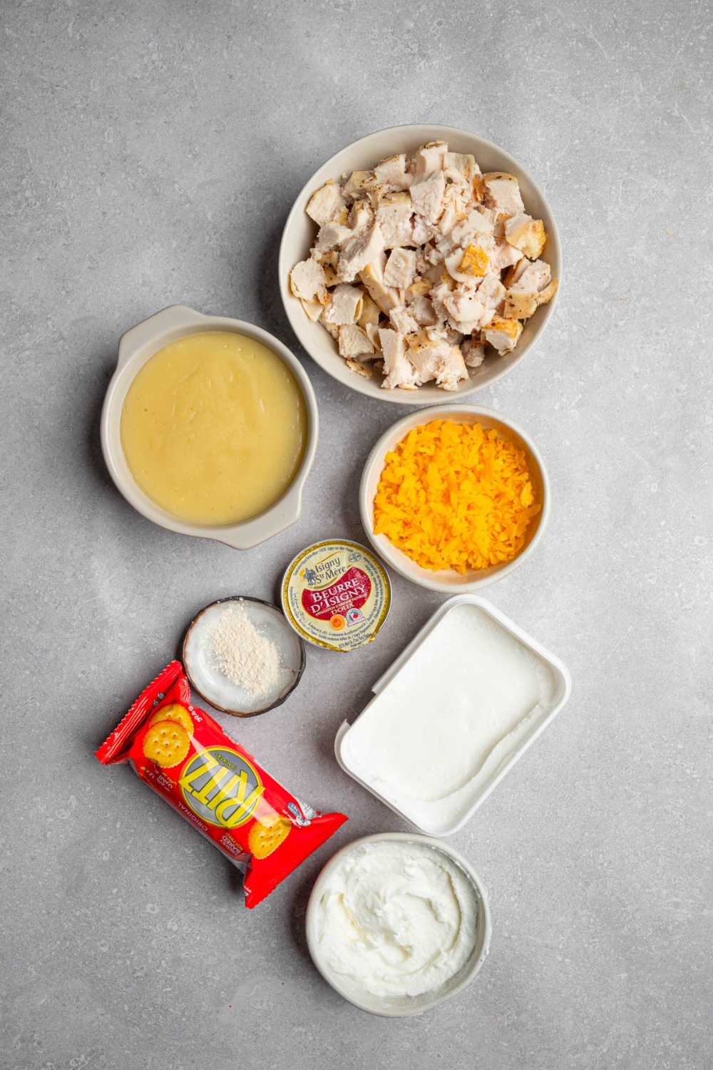 An overhead shot of several bowls in various sizes containing ingredients to make Neiman Marcus chicken casserole including cubed chicken, cream cheese, Ritz crackers, shredded cheddar cheese, sour cream, and cream of chicken soup.