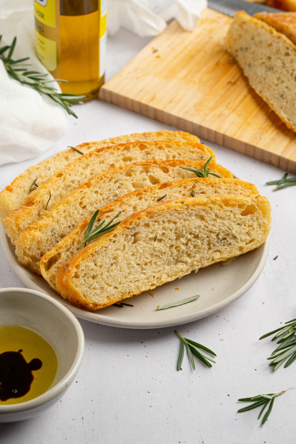Several slices of garlic rosemary bread on a white plate garnished with fresh rosemary. The plate is on a white counter with a wooden board of bread and jar of olive oil.