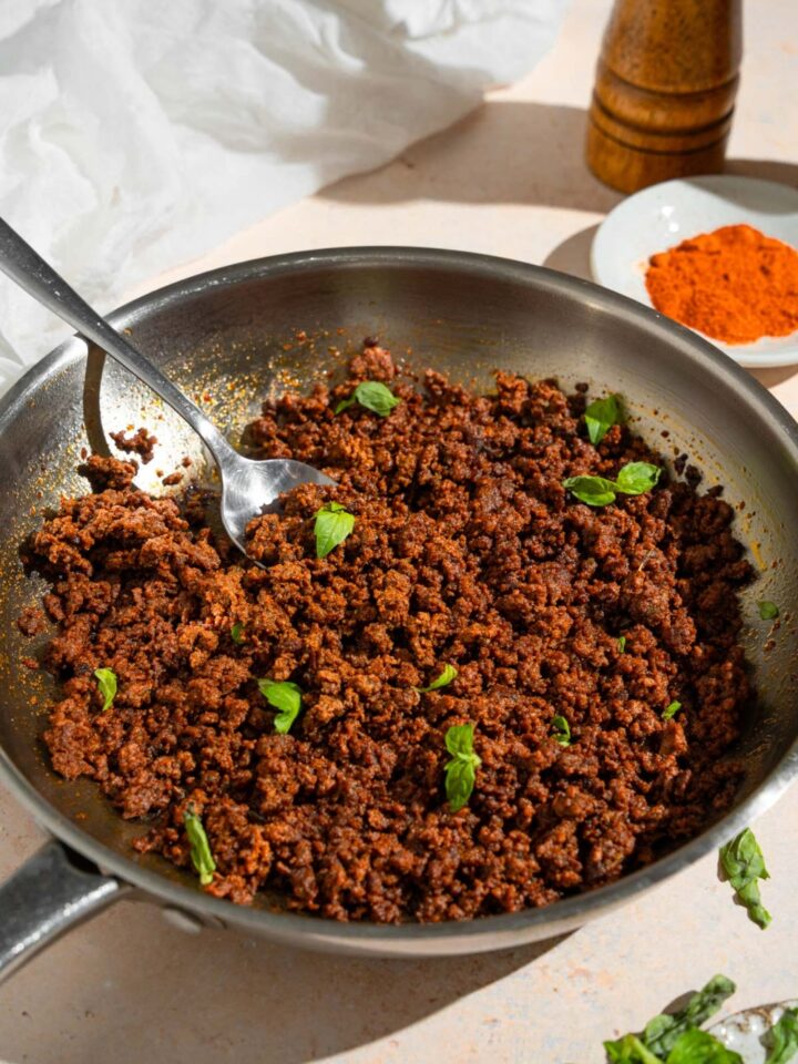 A skillet with cooked ground beef chorizo garnished with fresh basil. A fork is in the chorizo. The skillet is on a tan counter with a small plate of spices and white cloth napkin.