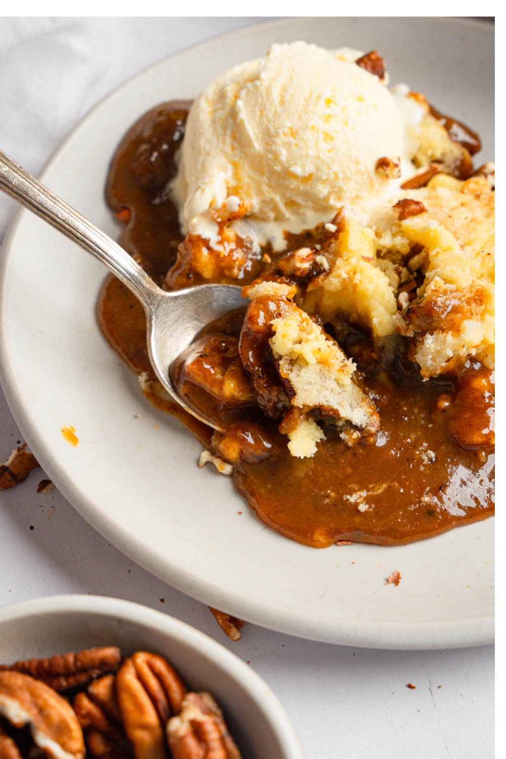 A slice of pecan pie dump cake topped with a scoop of vanilla ice cream on a white plate. There is a spoon taking a bite of the cake.