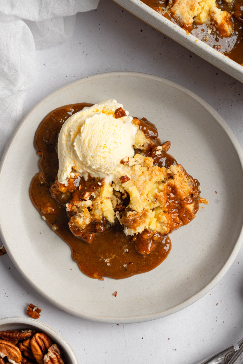 A slice of pecan pie dump cake topped with a scoop of vanilla ice cream on a white plate. The plate is on a white counter with a baking dish of cake and small bowl of pecans.