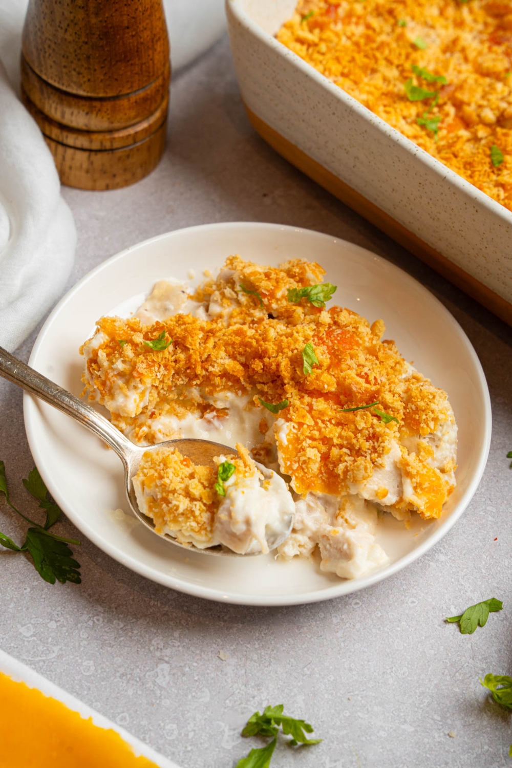 A plate of Neiman Marcus chicken casserole garnished with fresh parsley. A spoon is taking a bite of the casserole. The plate is on a white counter with a baking dish of casserole.