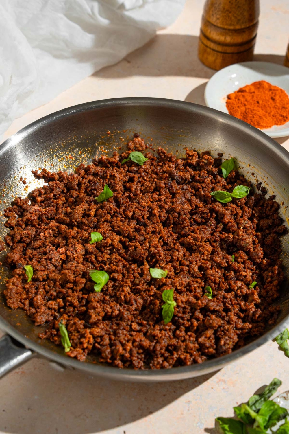 A skillet with cooked ground beef chorizo garnished with fresh basil. The skillet is on a tan counter with a small plate of spices and white cloth napkin.