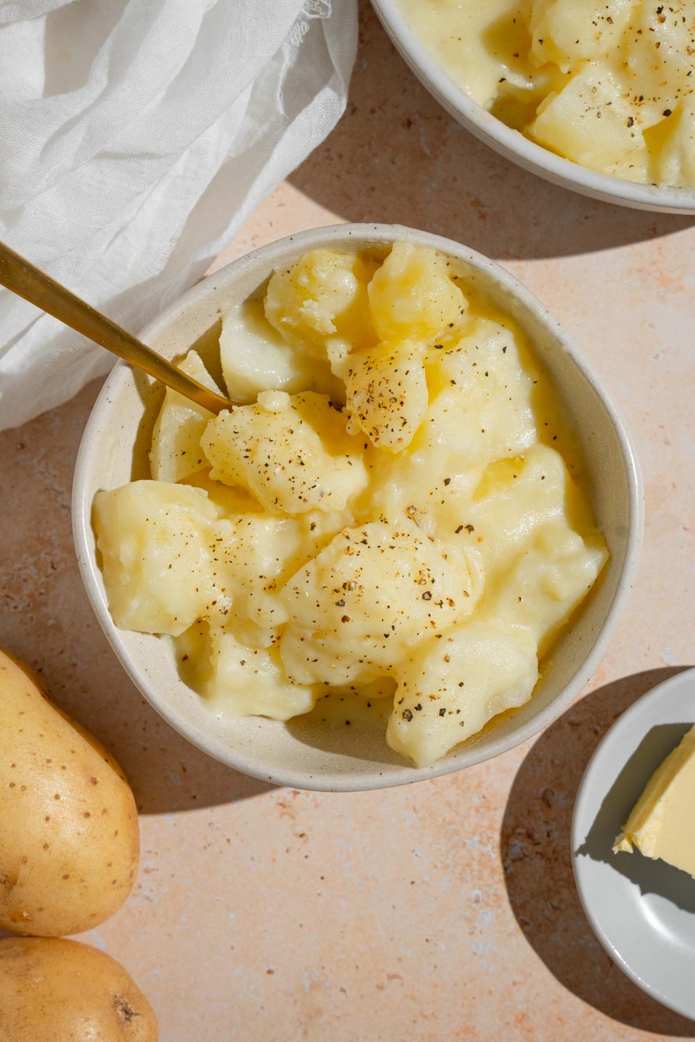 Stewed potatoes in a white bowl finished with ground pepper. There is a spoon in the bowl. The bowl is on a tan counter with an additional bowl of potatoes and a plate of butter.