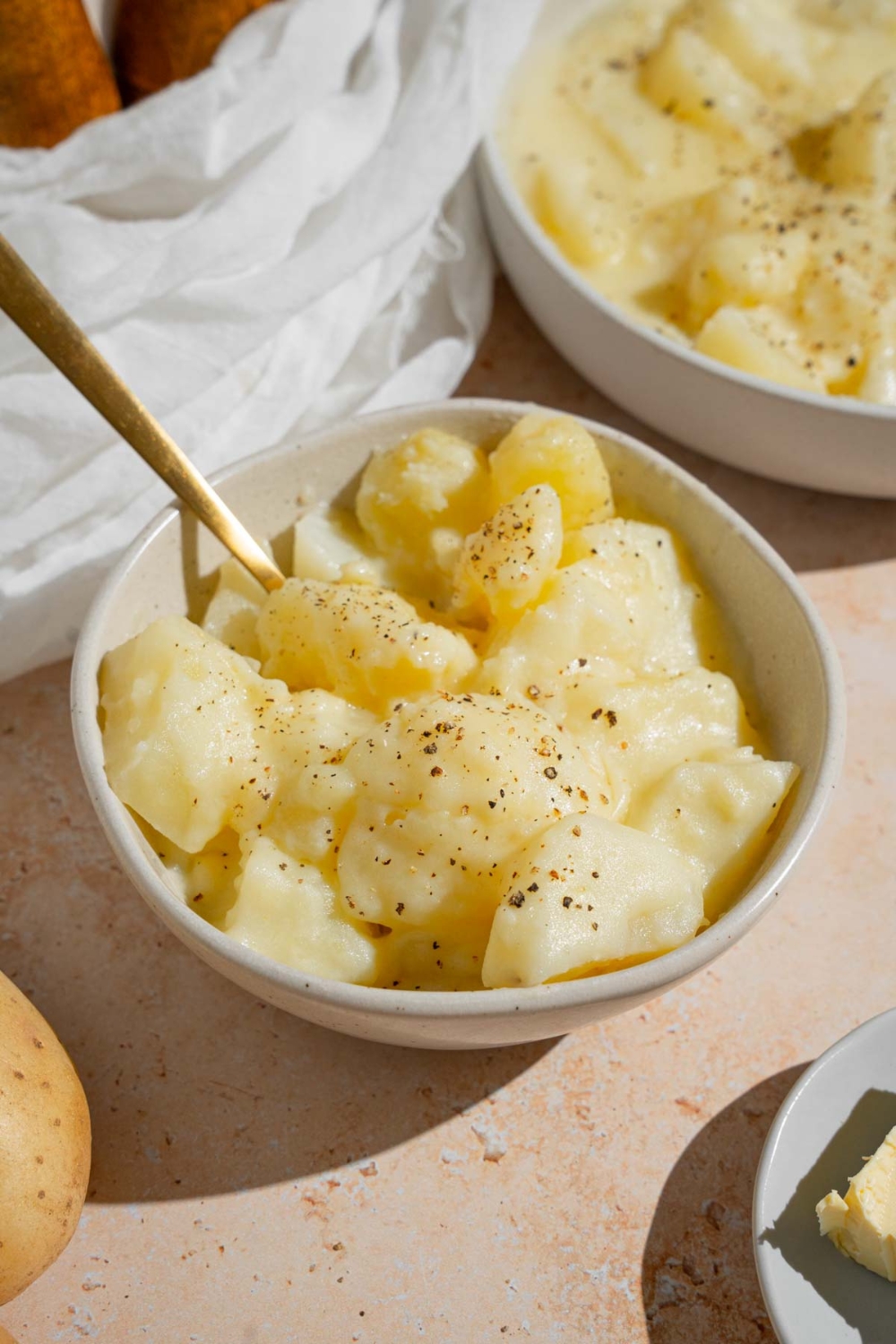 Stewed potatoes in a white bowl finished with ground pepper. There is a spoon in the bowl. The bowl is on a tan counter with an additional bowl of potatoes and a plate of butter.