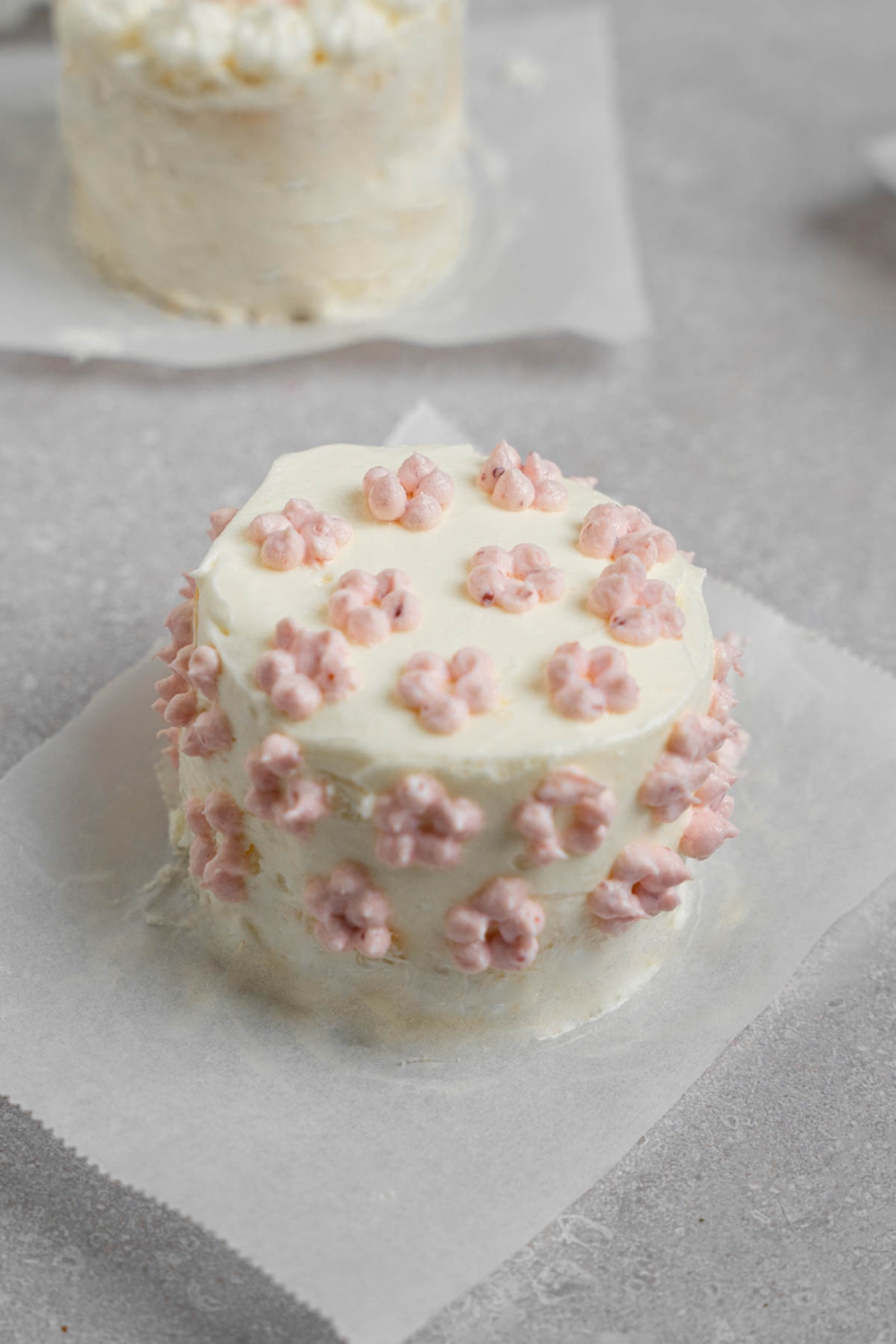 A vanilla lunch box cake with dotted purple icing on a piece of parchment paper. The plate is on a white counter with another cake.