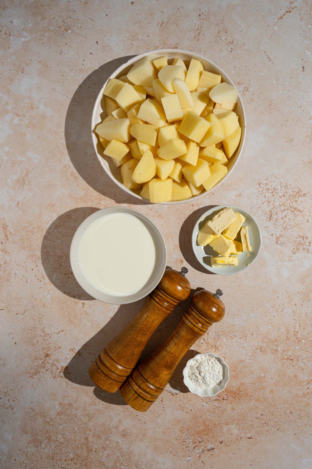An overhead shot of several bowls in various sizes containing ingredients to make stewed potatoes including cut potatoes, sliced butter, milk, flour, and pepper mills.