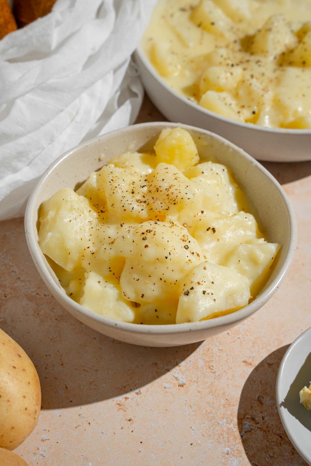 A bowl of stewed potatoes finished with ground pepper. The bowl is on a tan counter with an additional bowl of potatoes and whole potatoes.