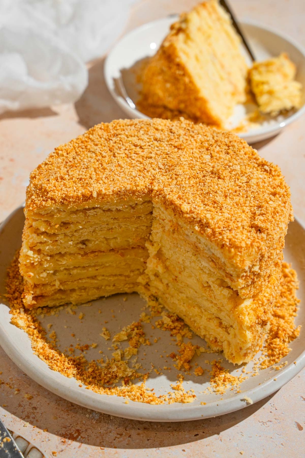 A Napoleon cake with a third of the slices removed. The cake is on a white plate. The plate is on a tan counter with a white cloth napkin and plate with a slice of cake.