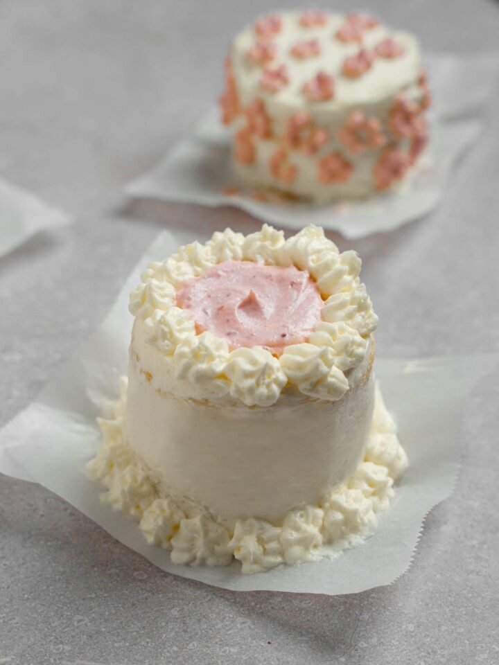 A vanilla lunch box cake with purple icing. The cake is on a piece of parchment paper on a white counter with other cakes.