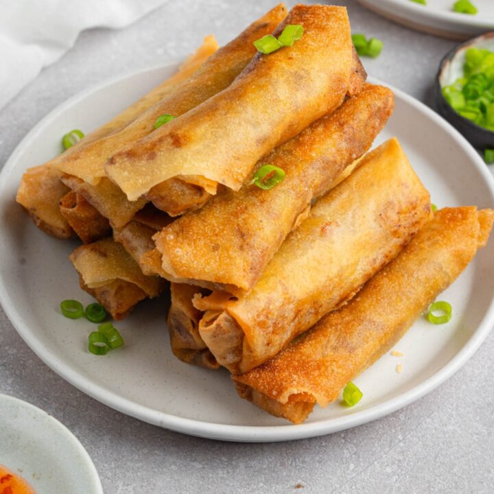 A white plate with a stack of vegetable egg rolls garnished with sliced green onion. The plate is on a white counter with a plate of garnishes, bowl of dipping sauce, and plate of egg rolls.