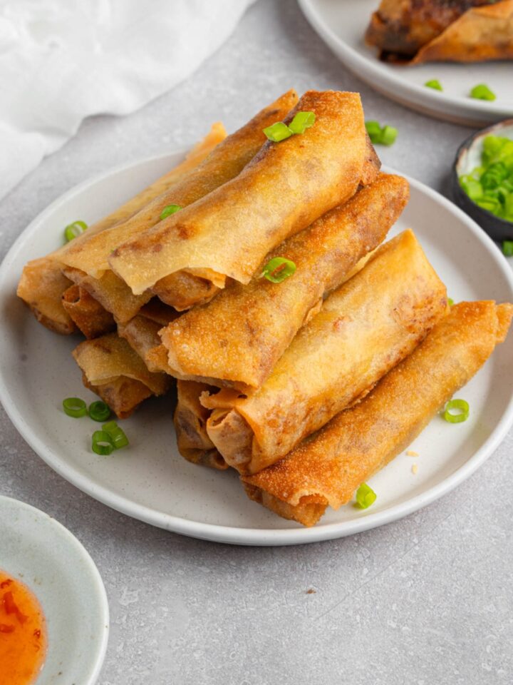 A white plate with a stack of vegetable egg rolls garnished with sliced green onion. The plate is on a white counter with a plate of garnishes, bowl of dipping sauce, and plate of egg rolls.