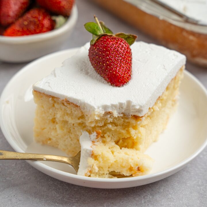 A white plate with a slice of milk cake topped with a fresh strawberry. There is a fork with a bite of cake on the plate. The plate is on a white counter with a tray of cake and bowl of strawberries.