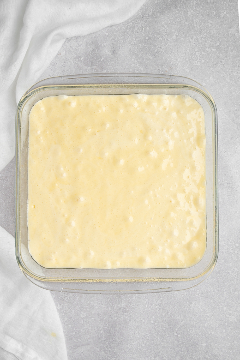 A glass baking dish with milk cake batter. The dish is on a white counter with a white cloth napkin.