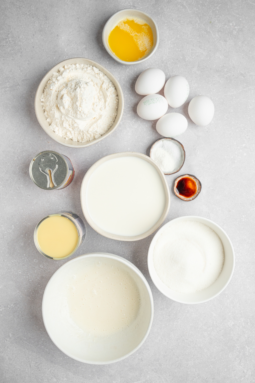 An overhead shot of several bowls in various sizes containing ingredients to make milk cake including eggs, flour, sugar, vanilla, milk, evaporated milk, condensed milk, and whipped cream.