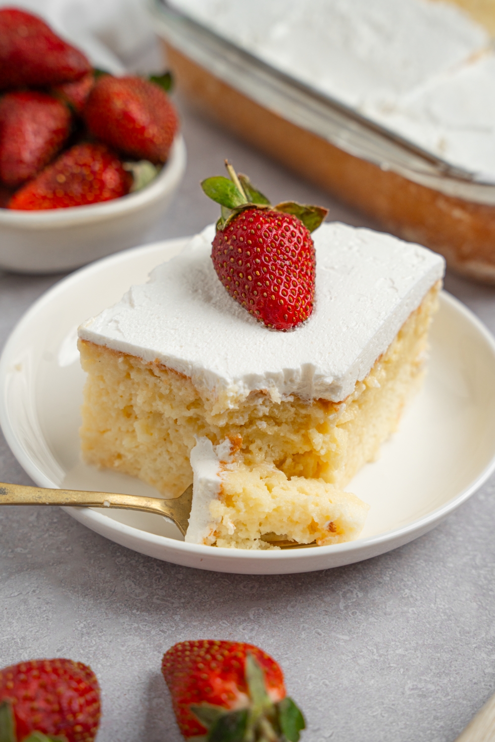 A white plate with a slice of milk cake topped with a fresh strawberry. There is a fork with a bite of cake on the plate. The plate is on a white counter with a tray of cake and bowl of strawberries.