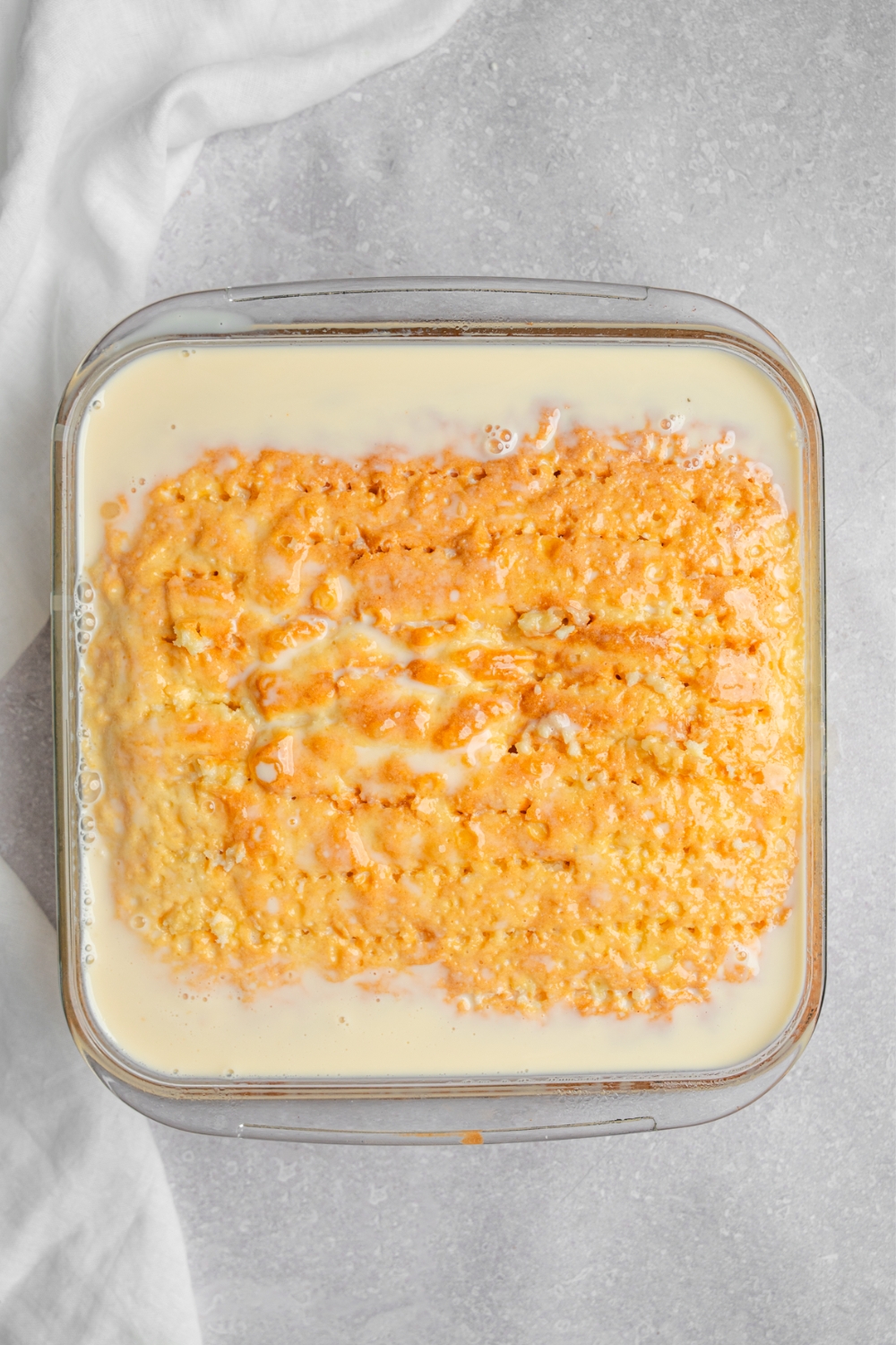 A square glass baking dish with milk cake sponge topped with a milk mixture. The dish is on a white counter with a white cloth napkin.