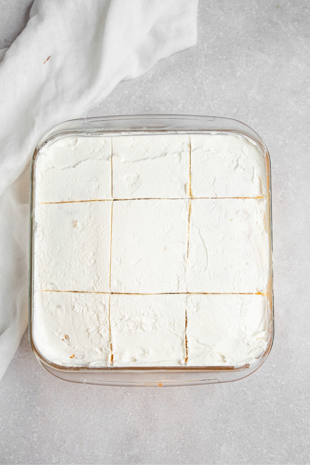 A square glass baking dish with milk cake topped with whipped cream. The cake is sliced into 9 slices. The dish is on a white counter with a white cloth napkin.
