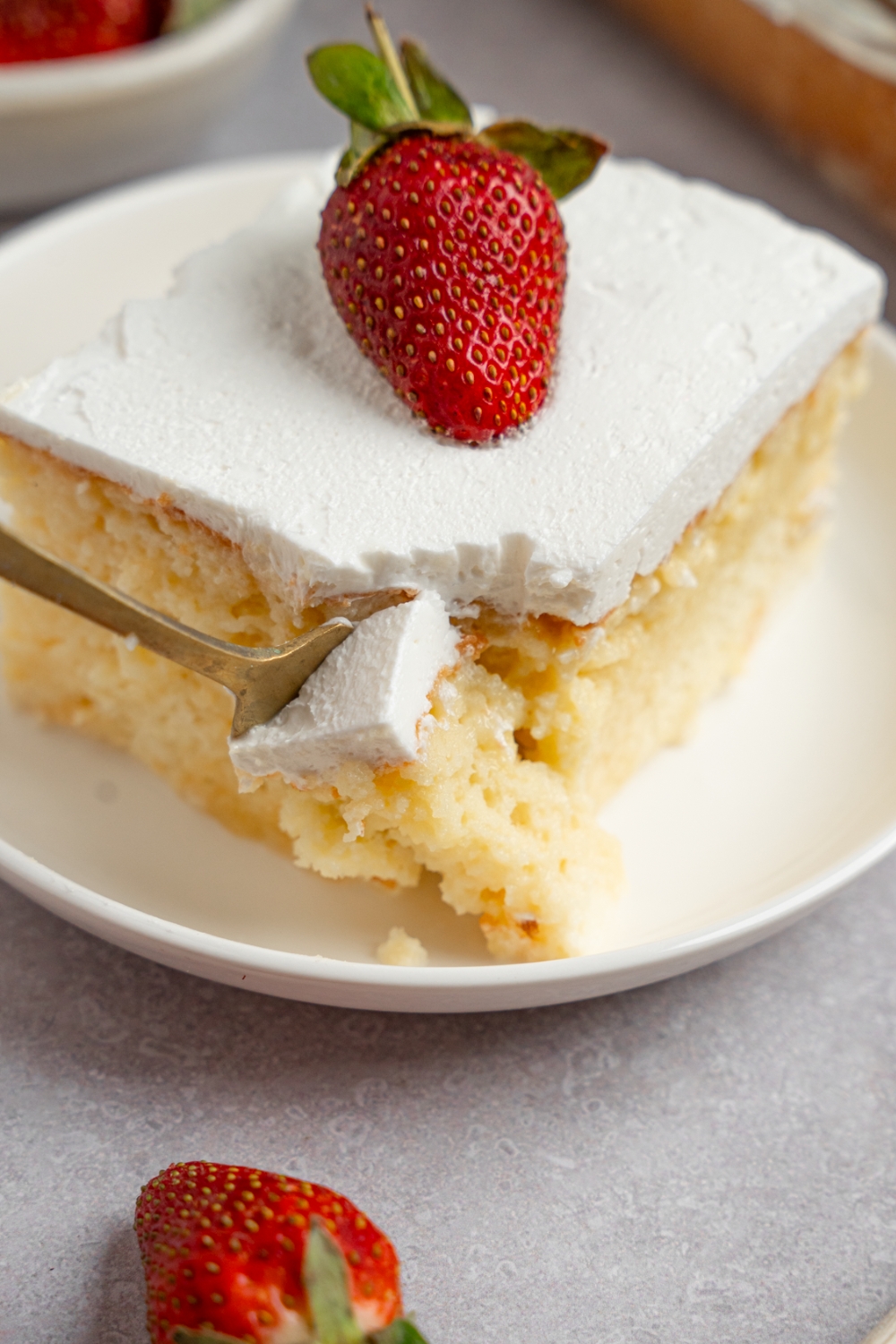 A slice of milk cake topped with a strawberry on a white plate. A fork is taking a bite from the slice. The plate is on a white counter.