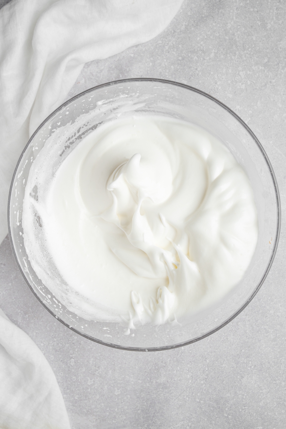 A glass bowl with beaten egg whites. The bowl is on a white counter with a white cloth napkin.