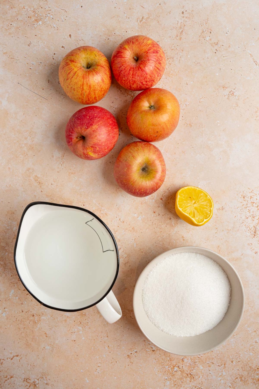 An overhead shot of ingredients to make homemade apple jelly including several apples, lemon, sugar, and water.