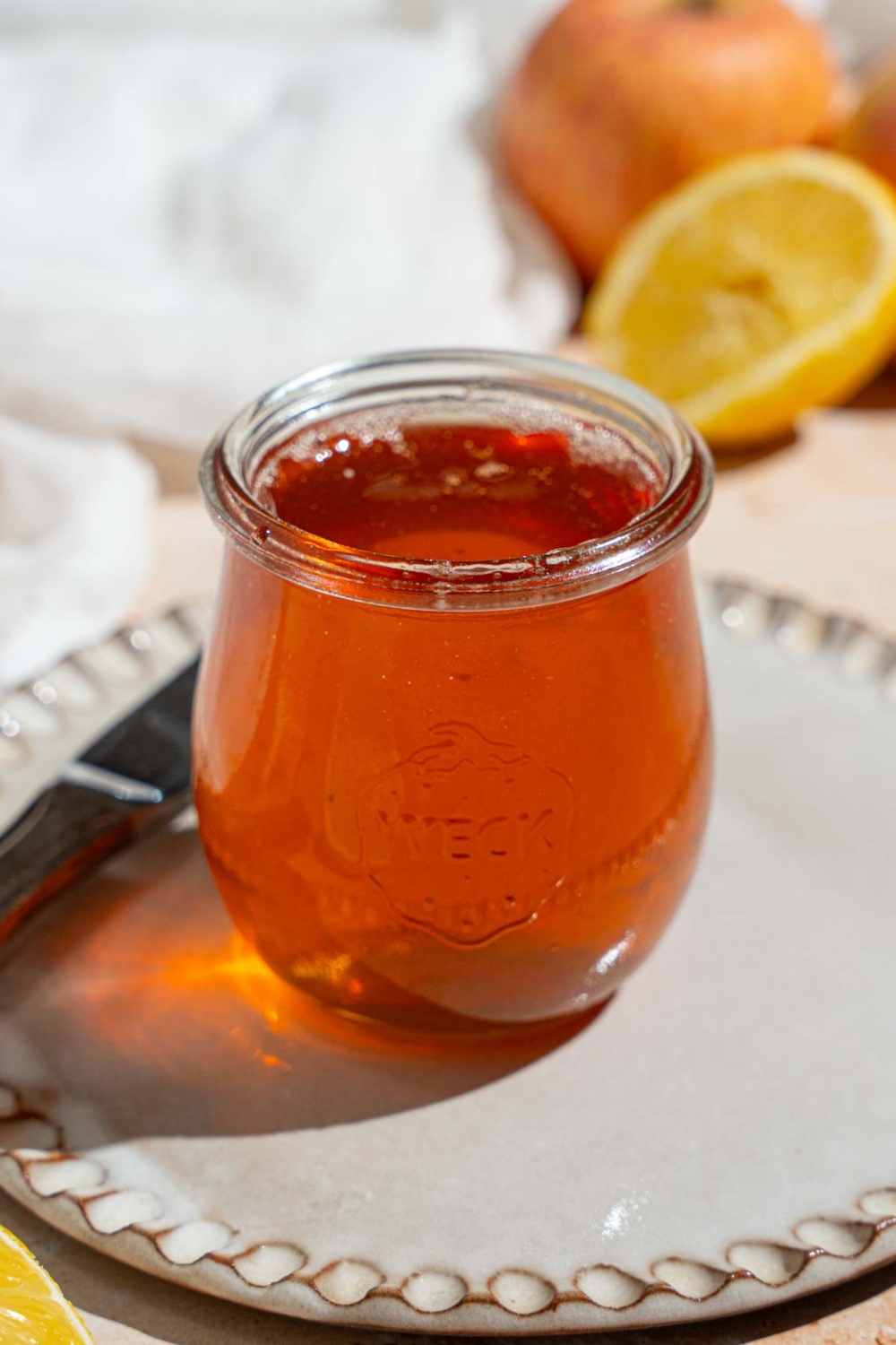 A small glass jar with homemade apple jelly. The jar is on a ceramic plate with a knife. The plate is on a tan counter with sliced lemon, apples, and a white cloth napkin.