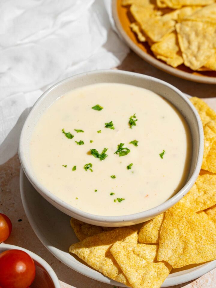 A bowl of Chipotle queso garnished with chopped parsley. The bowl is on a plate with tortilla chips. The plate is on a tan counter with a bowl of tortilla chips and white cloth napkin.