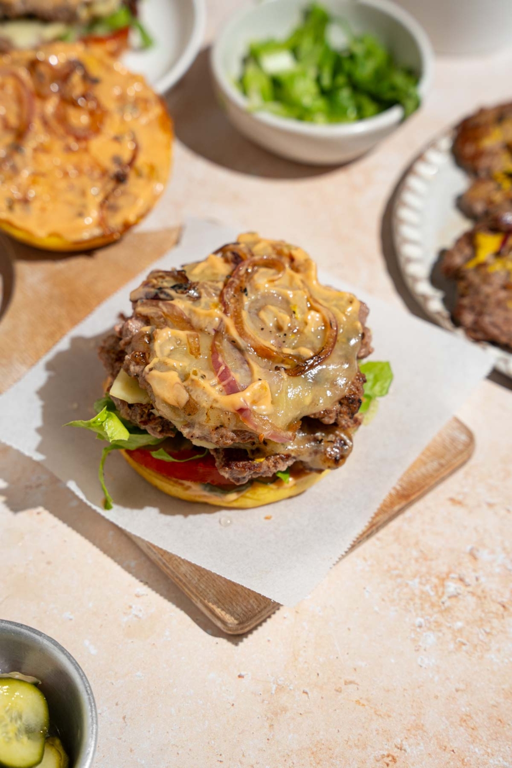 An open copycat In-N-Out burger. The burger is on a wooden board lined with parchment paper. The board is on a tan counter with a plate of burger patties and bowl of shredded lettuce.