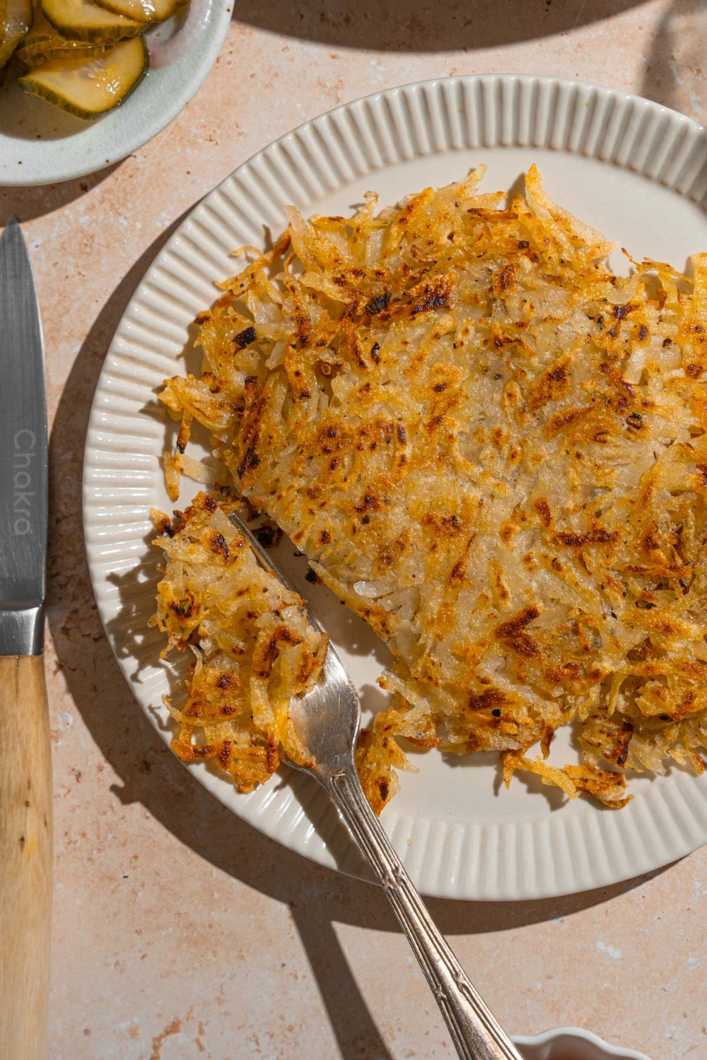 A white plate with copycat Waffle House hash browns. There is a fork taking a bite of hash browns. The plate is on a tan counter with a knife.