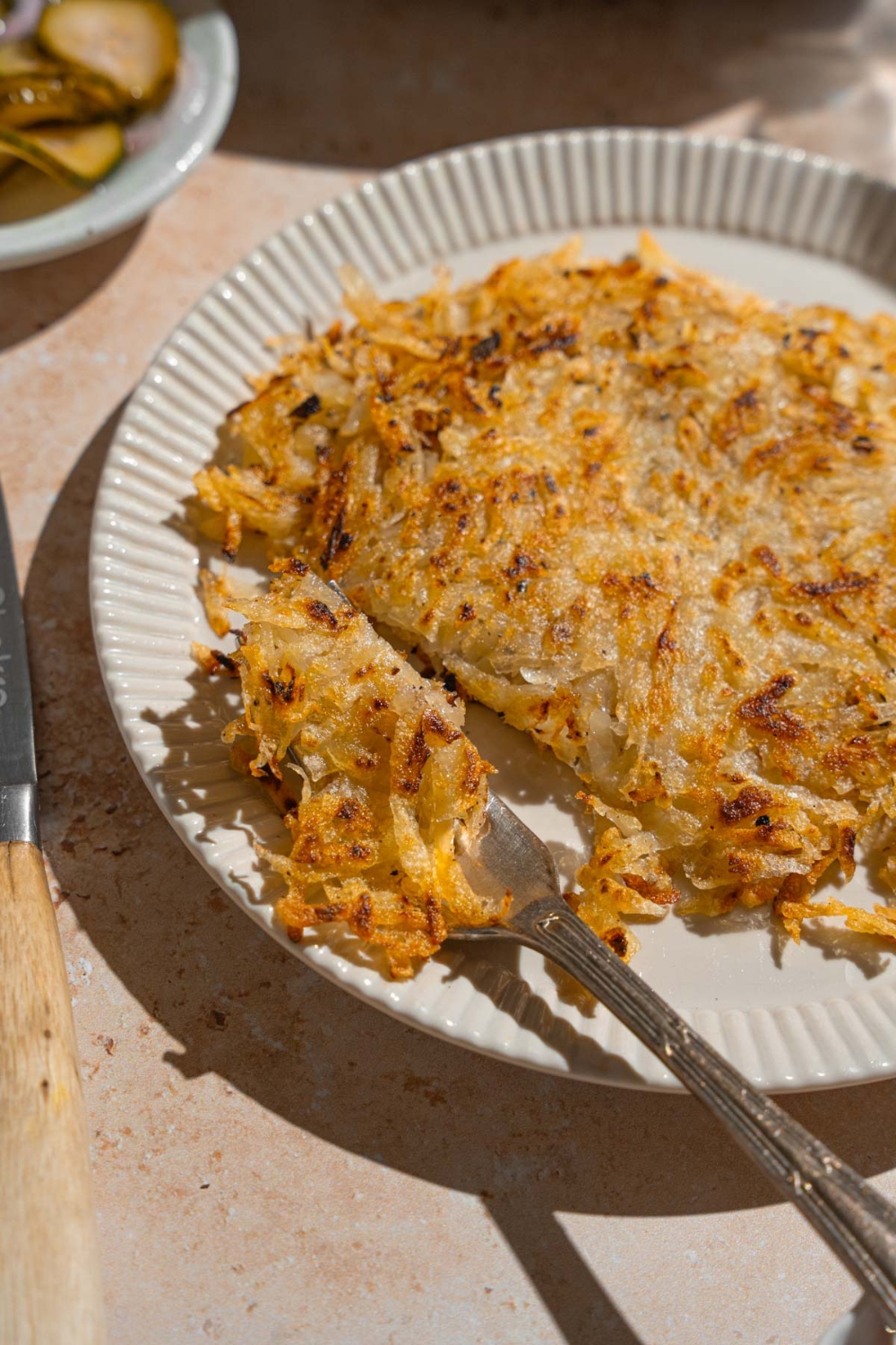 A white plate with copycat Waffle House hash browns. There is a fork taking a bite of hash browns. The plate is on a tan counter with a knife.