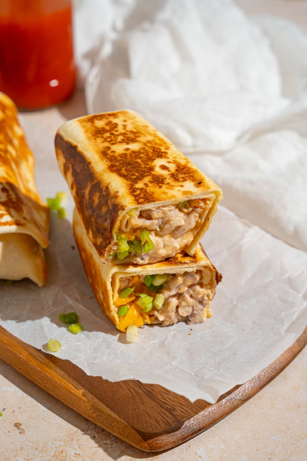 A cheesy garlic chicken wrap sliced in half on a wooden board lined with parchment paper. There is an additional wrap on the board. The board is on a tan counter with a white cloth napkin.