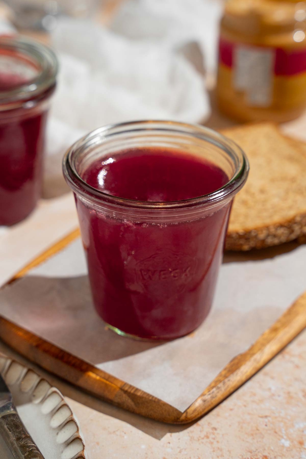A glass jar with chokecherry pudding. The jar is on a wooden board lined with parchment paper with a slice of bread. The board is on a tan counter with a white cloth napkin and additional jar of pudding.