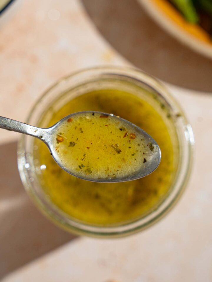 A close up of a spoon with copycat Texas Roadhouse italian dressing. There is a jar of dressing on a tan counter blurred in the background.