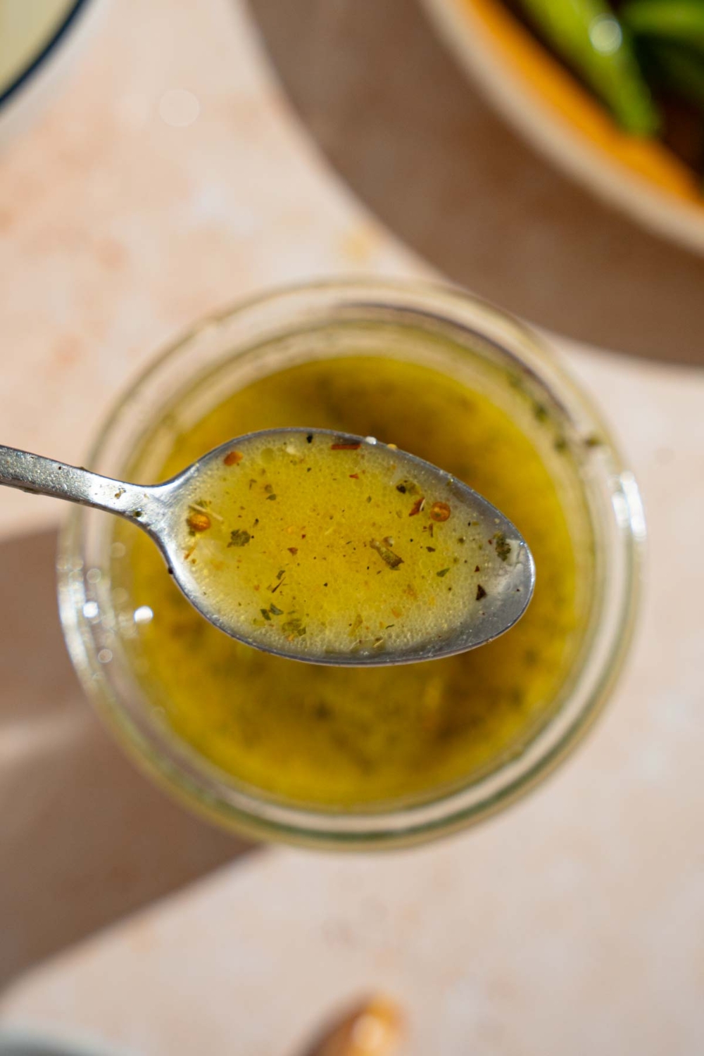 A close up of a spoon with copycat Texas Roadhouse italian dressing. There is a jar of dressing on a tan counter blurred in the background.