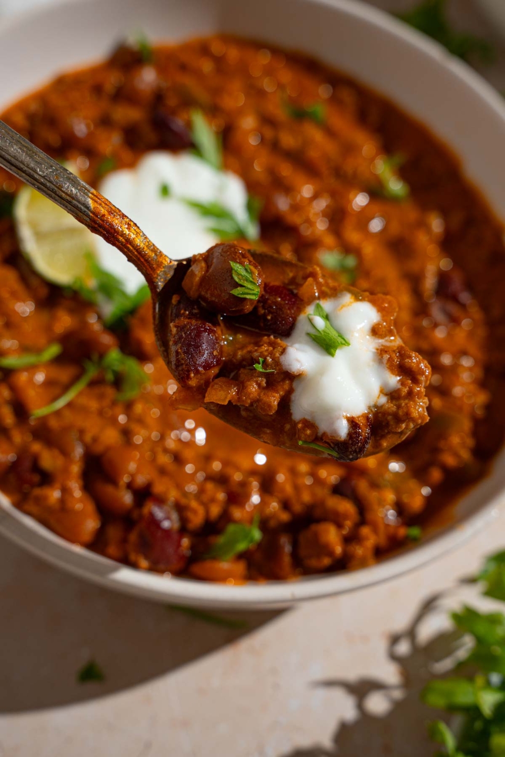 A close up of a spoon with a bite of copycat Zippy's chili garnished with sour cream and fresh parsley. There is a bowl of chili blurred in the background.